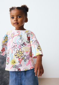 Chemise à manches courtes et colorée présentant une variété de fleurs dessinées, d'étoiles et de gribouillis vibrants sur un fond rose clair.