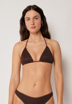 Frau mit langen dunklen Haaren, die ein dunkelbraunes, glitzerndes Triangel-Bikinitop und einen passenden Bikinihöschen trägt, steht vor einem schlichten Hintergrund.