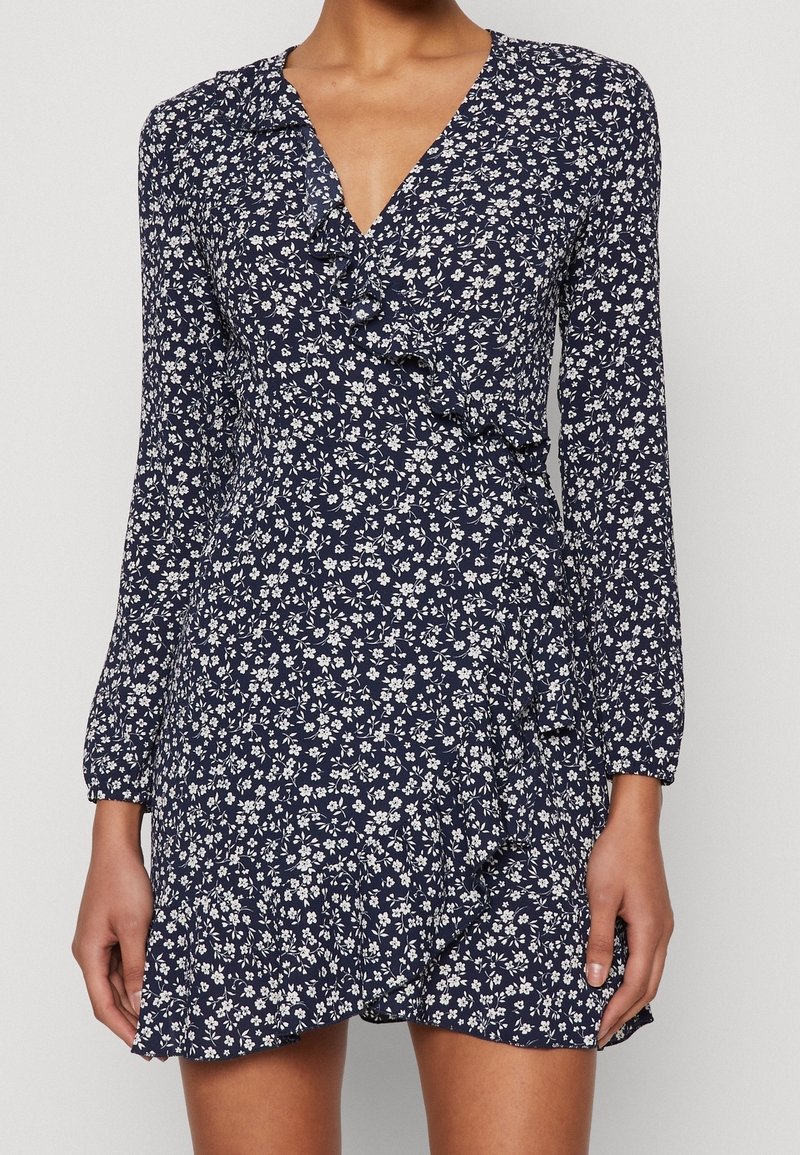 Femme portant une robe portefeuille bleu marine à manches longues avec imprimé floral blanc et bordure à volants, photographiée du cou jusqu'à mi-cuisse sur un fond uni.
