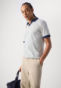 Polo rayé en beige clair avec des accents bleu marine, présentant un col et des manches courtes. Porté avec un pantalon clair et tenant un sac sombre.