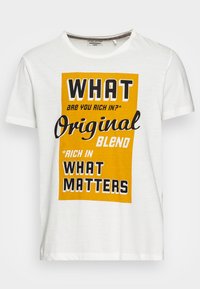 Valge puuvillane t-särk, millel on julged musta ja oranži värvi tekstid sõnadega nagu "MIS", "Original Blend" ja "MIS ON OLULINE." Lühikesed varrukad, ümar kaelus.