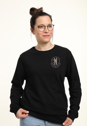 Donna con occhiali e bun, che indossa un felpa nera con un emblemma grigio chiaro sul petto, mentre tiene il bordo e sorride leggermente, su uno sfondo chiaro.