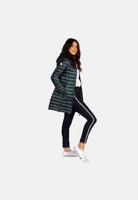 Doudoune vert foncé avec des coutures horizontales, une capuche, des poches zippées, portée sur un haut blanc, associée à un jean foncé avec des rayures blanches.