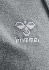 Tejido de algodón gris con un logo bordado de diseño simétrico y la palabra "hummel" en hilo blanco.