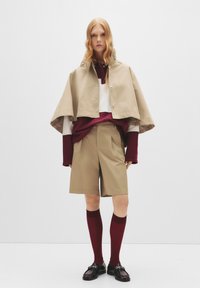 Giacca a maniche corte beige, camicia a maniche lunghe bordeaux, pantaloni beige, calze al ginocchio bordeaux e scarpe nere. Tessuto morbido, dettagli minimi.