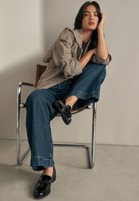 Une personne assise sur une chaise en métal, portant un blazer beige à carreaux, un haut marron foncé, un jean large bleu et des mocassins noirs brillants avec un détail nœud.