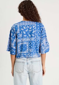 Blouse courte bleue avec des motifs paisley et floraux blancs, manches amples et col rond, fabriquée en tissu léger.