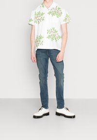 Bílé tričko s krátkými rukávy a zeleno-žlutými květinovými vzory, doplněné modrými džínami a bílými botami s černými podrážkami a šňůrkami.