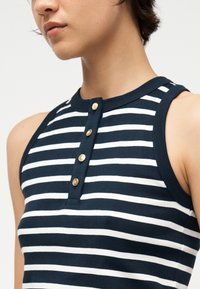 Κοντινό πλάνο ενός ατόμου που φοράει αμάνικη μπλούζα με ναυτικό μπλε και λευκές ρίγες, με τέσσερα χρυσά κουμπιά στην εμπρόσθια πλακέτα.