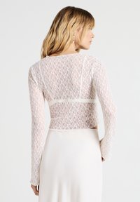 Top corto bianco in pizzo con maniche lunghe, caratterizzato da un delicato motivo floreale e una scollatura rotonda, indossato su una gonna liscia di colore chiaro.