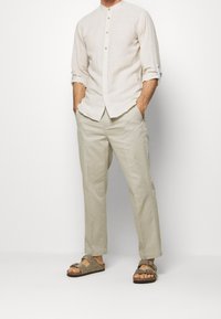 Camisa de lino beige claro con cuello mao, botonadura al frente y mangas enrolladas, combinada con pantalones caqui claros y sandalias marrones.