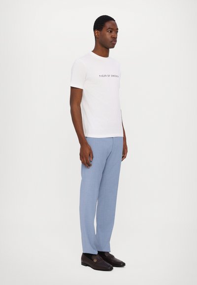 Weißes T-Shirt mit kurzen Ärmeln und dem Schriftzug "TIGER OF SWEDEN", kombiniert mit hellblauen Hosen und dunkelbraunen Mokassins mit goldenen Akzenten.