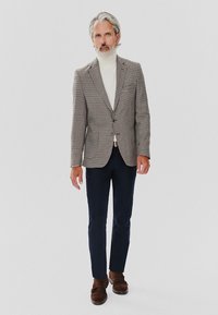 Marynarka w deseń houndstooth w odcieniach brązu, biała golf, granatowe spodnie i brązowe zamszowe buty na neutralnym tle.