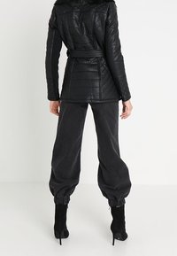 Chaqueta negra de cuero acolchada con cuello de piel sintética, cintura con cinturón y mangas largas; combinada con pantalones sueltos gris oscuro y botas negras de tacón.