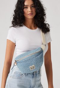Marsupio in tessuto intrecciato azzurro chiaro con superficie testurizzata, zip in metallo argentato e tracolla regolabile beige. Presenta un'etichetta sul davanti.