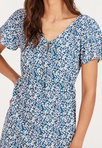 Robe fleurie bleue avec manches courtes, col en V avec détail à nouer et taille cintrée. Le tissu a une texture douce avec de petites fleurs roses et blanches.