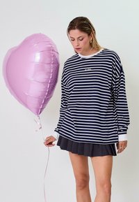 Tengerés kék és fehér csíkos, hosszú ujjú póló, kerek nyakkivágással, sötét plissé szoknyával párosítva, egy rózsaszín, szív alakú lufit tartva.