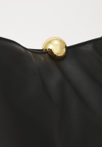 Crna kožna torba s sjajnim zlatnim okruglim kopčom sa ugraviranim natpisom "Furla". Tekstura je glatka i elegantna, ističući moderni dizajn.