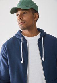 En ung mann iført en grønn caps, en marineblå zip-up hettegenser og en hvit t-skjorte som ser til siden mot en enkel lys bakgrunn.