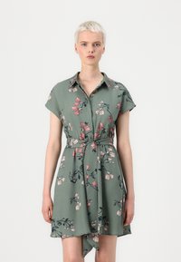 Abito floreale verde con maniche corte, colletto e vita con fiocco. Presenta un motivo di fiori rosa e blu scuro e un orlo che si drappeggia in modo asimmetrico.