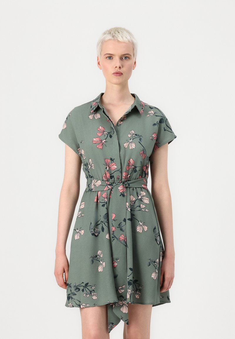 Abito floreale verde con maniche corte, colletto e vita con fiocco. Presenta un motivo di fiori rosa e blu scuro e un orlo che si drappeggia in modo asimmetrico.