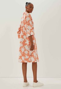 Bloemig shirtjurk in perzik met witte bloemenpatronen, kraag, driekwart mouwen en een knie-lange snit. Draagt een paar witte sneakers.