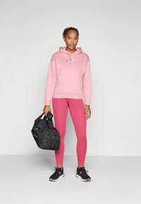 Camisola com capuz cor-de-rosa com logótipo, leggings cor-de-rosa a condizer, sapatilhas pretas e um saco desportivo preto. Tecido macio e corte descontraído.