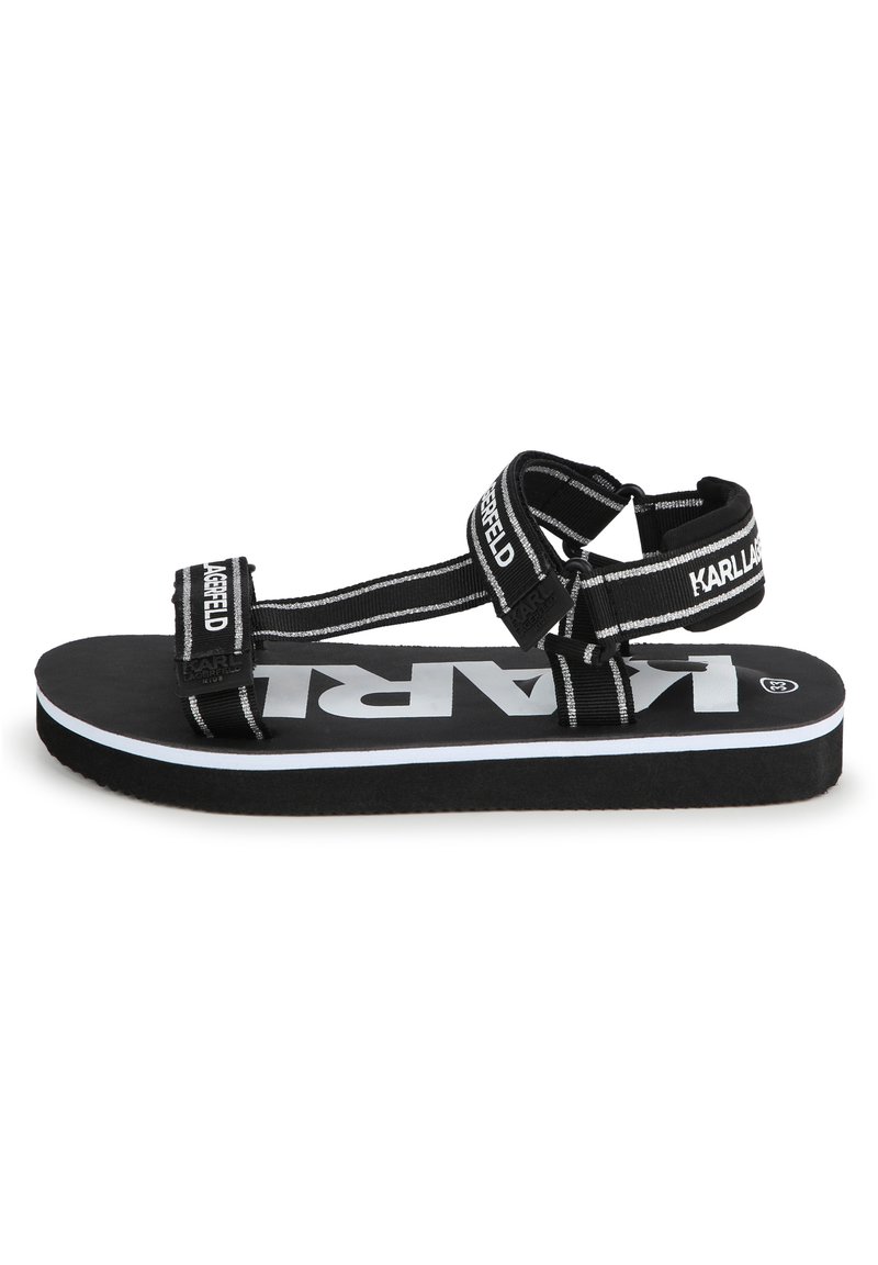 Sandales noires avec une large bride présentant une inscription blanche. Semelle douce avec une bordure blanche, brides ajustables et conception plate pour le confort.