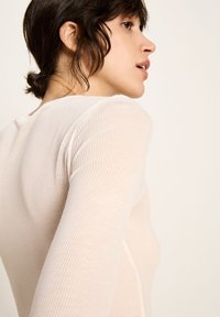 Junge Frau mit dunklem Haar, das zu einem niedrigen Dutt frisiert ist, trägt ein geripptes, beige Oberteil mit langen Ärmeln und schaut nach rechts vor einem schlichten Hintergrund.