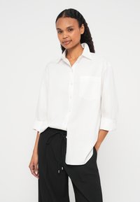 Valge nööbitav triiksärk terava krae ja ühe rinnataskuga, sobitatud mustade pükstega, millel on paelaga vöökoht ja lõdvestunud lõige.