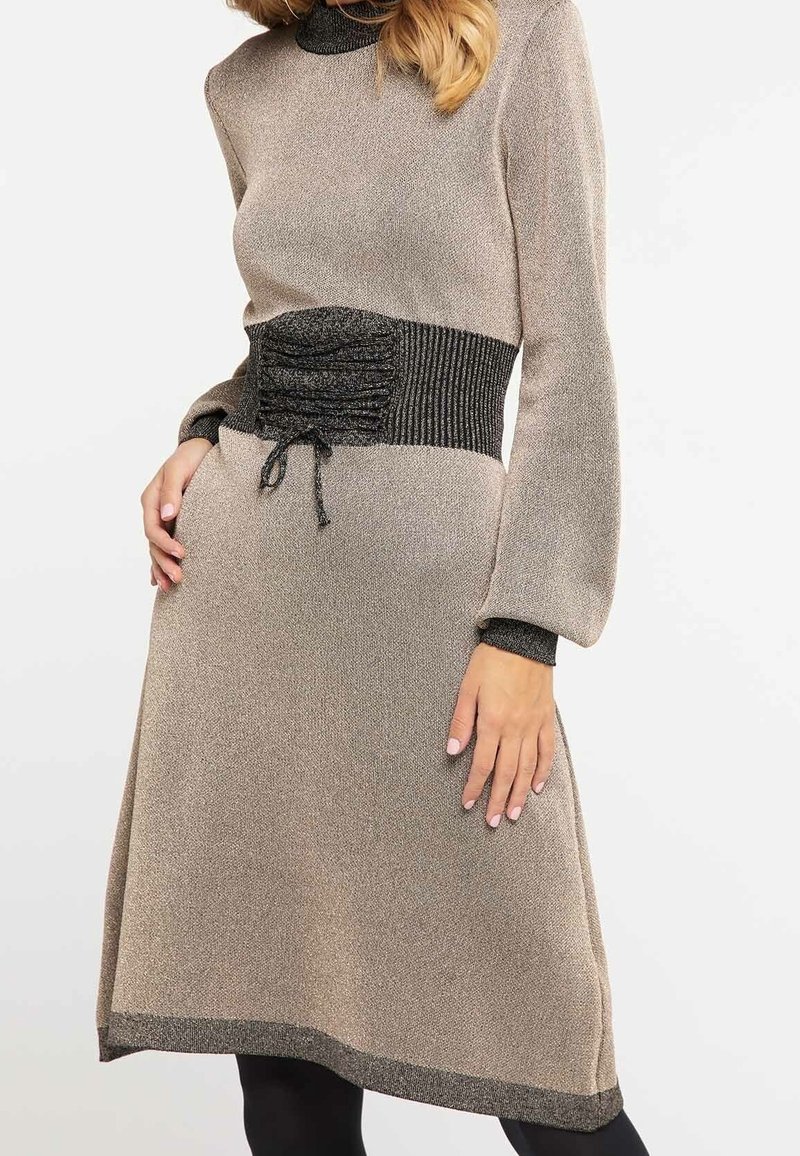 Robe midi en tricot beige clair avec une taille noire texturée et moulante, des manches longues et des accents noirs.