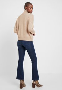 Bež pleten pulover iz mešanice volne z ovratnikom, sproščen kroj, rebrasti manšeti in spodnji rob, kombiniran s temno modrimi razširjenimi kavbojkami in škornji z visokimi petami v vzorcu kačje kože.