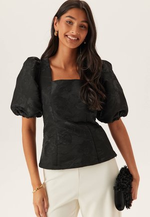 Frau lächelt, trägt schwarzen Jacquard-Puffärmel-Top mit eckigem Ausschnitt, weiße Hose, goldene Creolen, Armband und hält eine schwarze Blumen-Clutch.