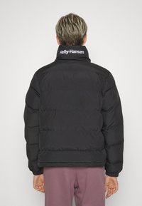 Svart pufferjacka med hög krage, med synliga sömmar, en logga på nacken och en åtsittande passform. Bärs med rosa byxor.