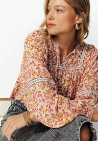 Femme portant un blouse fleuri rose et orange avec des manches bouffantes, des boucles d'oreilles en forme de goutte d'or, un collier fin et un bracelet en perles d'or.