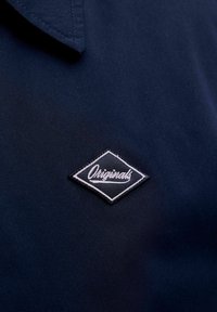 Tissu bleu foncé avec un patch en forme de losange portant le mot « Originals » en couture cursive blanche près du bord du col.