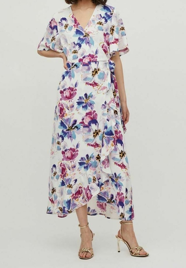 Femme portant une robe portefeuille blanche avec des motifs floraux bleus, violets et roses, des manches flottantes et des sandales à talons hauts dorées.