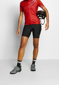 Czerwony trykot kolarski z przednim zamkiem i gradientowym wzorem kropek; czarne spodenki kolarskie; szare buty kolarskie; czarny kask trzymany w ręku.