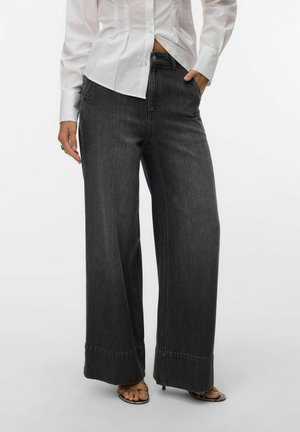 Femme portant un jean noir à jambes larges, une chemise blanche boutonnée et des sandales à talons ouvertes, debout avec une main dans la poche.