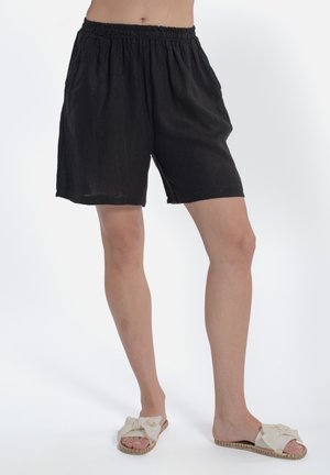 Kobieta w luźnych czarnych szortach i kremowych sandałach wsuwanych z kokardkami, stojąca na jednolitym białym tle.