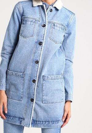 Jas z jasnego niebieskiego denimu z zapięciem na guziki, dwoma kieszeniami na piersi i dwoma dużymi kieszeniami z przodu, noszona z pasującymi spodniami jeansowymi.