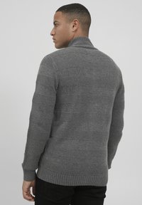 Grau gestrickter Pullover mit einem strukturierten Muster, hoher Kragen und gerippten Bündchen. Das Strickmuster zeigt unterschiedlich breite horizontale Streifen.