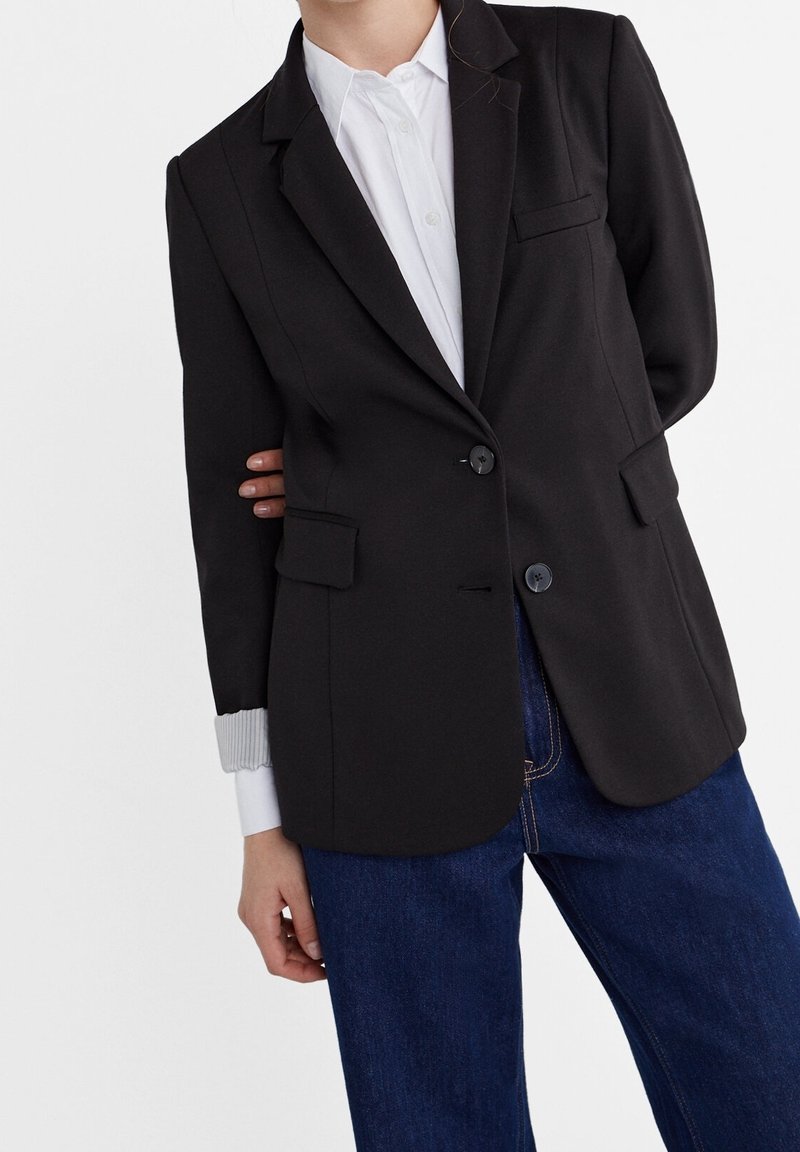 Personne portant un blazer noir à simple boutonnage sur une chemise blanche boutonnée et un jean bleu, debout devant un fond uni.