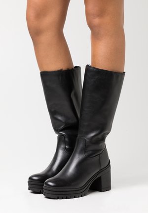 Bottes en cuir noires montantes jusqu'aux genoux avec talons carrés épais, portées sur des jambes nues, sur un fond blanc uni.