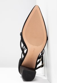 Chaussure à talon haut en daim noir avec bout pointu et semelle texturée. Présente des brides croisées et des accents en métal doré.
