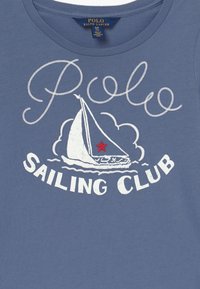 Blå bomullströja med vit skrift "Polo" och "Sailing Club", tillsammans med en design av en segelbåt och en röd stjärna som accent.