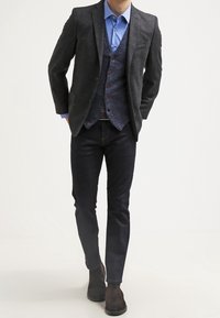 Grå texturerad kavaj över en blå skjorta och en marinväst, i kombination med mörka jeans och bruna läderstövlar. Vesten har dekorerade knappar.