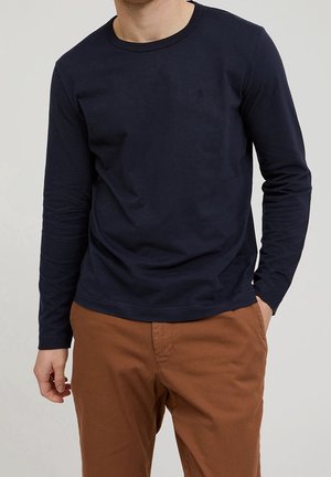 Homme portant une chemise à manches longues bleu marine et un pantalon marron, debout avec une main dans la poche devant un fond uni.