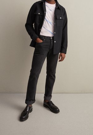 Mann trägt schwarze Jacke mit Knöpfen über weißem, gemustertem T-Shirt, schwarze hochgekrempelte Jeans und schwarze Lederschuhe, steht vor einer schlichten Wand.