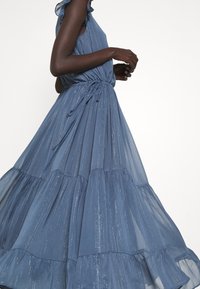 Blauwe maxi-jurk met lagen, gemaakt van lichtgewicht, zijdeachtig materiaal met een gestructureerde glans. Heeft een getailleerde bodice en ruffles op de schouderaccenten.